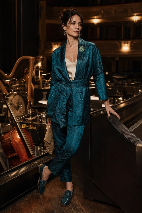 A woman wears a teal patterned wrap jacket with matching tapered trousers, a cream camisole, teal loafers, statement earrings, and a textured clutch.