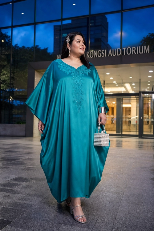 A woman wears a flowing teal kaftan dress with embroidered detailing, silver accessories, a beaded handbag, and strappy sandals outside an auditorium.