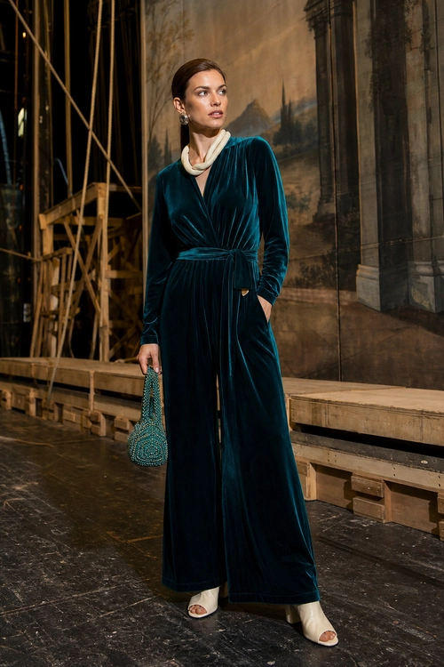A person wears a long-sleeved teal velvet wide-leg jumpsuit with a cream neck accessory, white sandals, and a beaded handbag.