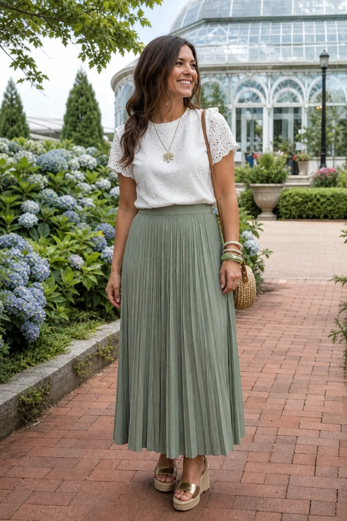 White Blouse and Sage Pleated Skirt Outfit Woman wearing a white short-sleeve eyelet blouse with a sage green pleated midi skirt, wedge sandals, and a woven shoulder bag.