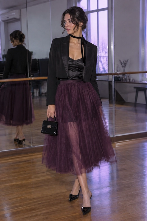 A woman wears a cropped black blazer, satin camisole, plum tulle midi skirt, pointed heels, and carries a small black handbag.