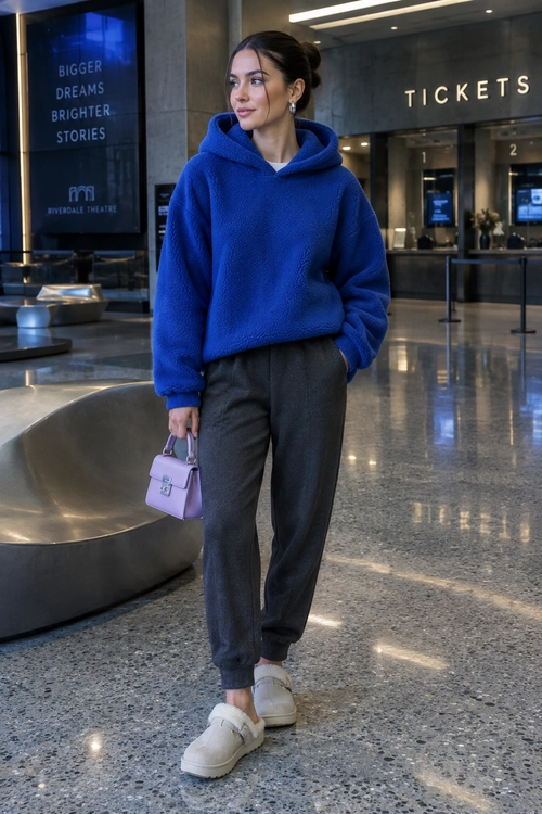 Person wearing a cobalt blue fleece hoodie, charcoal gray trousers, white slip-on sneakers, and carrying a lavender handbag.
