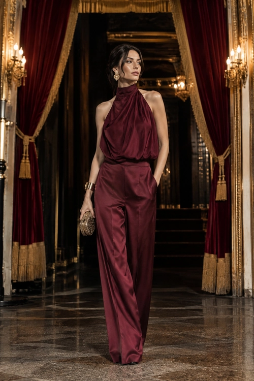 A woman wearing a burgundy draped halter jumpsuit with wide-leg trousers, gold earrings, a bracelet, and a beaded clutch.