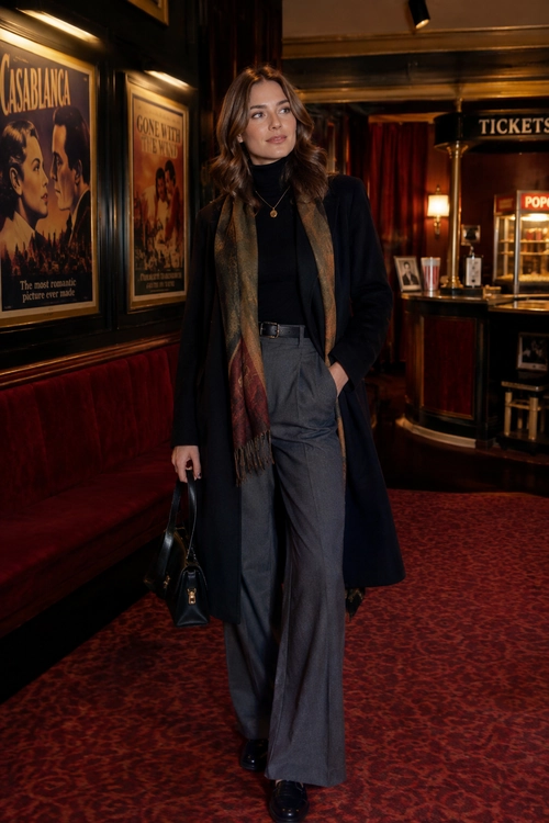 A woman wears a long dark coat, black turtleneck, patterned scarf, gray wide-leg trousers, and carries a black handbag in a theater.