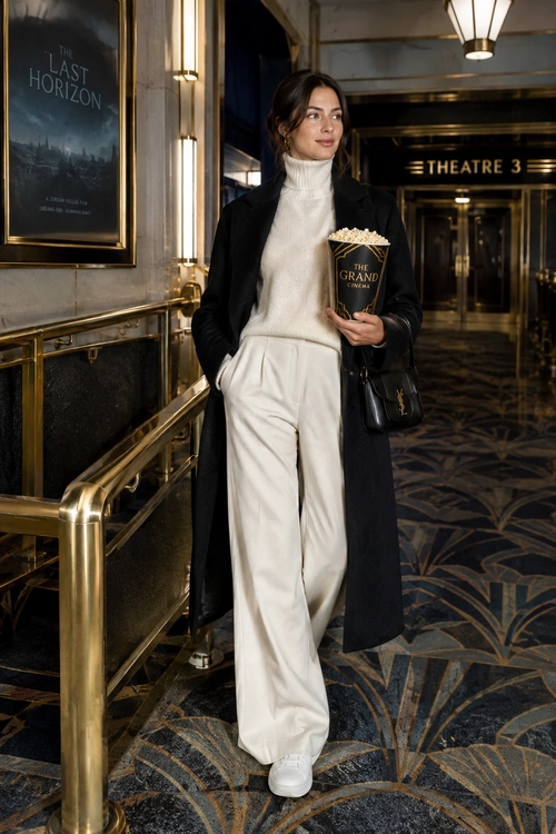 A woman wears a cream turtleneck sweater and wide-leg trousers with a long black coat and white sneakers at a cinema.