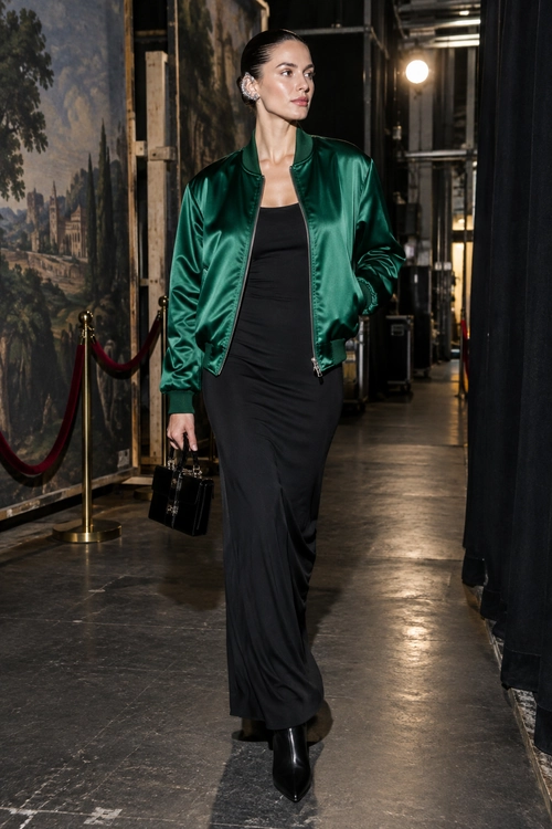 Emerald Bomber Jacket with Black Dress A woman wears an emerald green satin bomber jacket over a long black dress with pointed black boots and a structured black handbag.