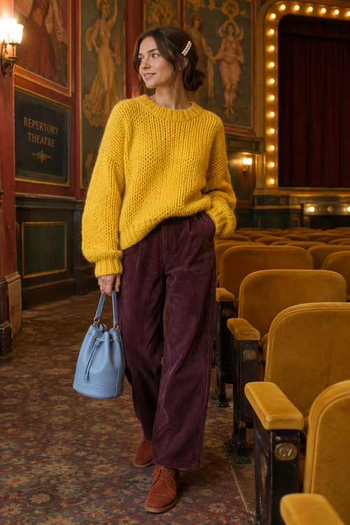 A woman wears a mustard yellow chunky knit sweater with burgundy wide-leg trousers, brown ankle boots, and a light blue handbag.