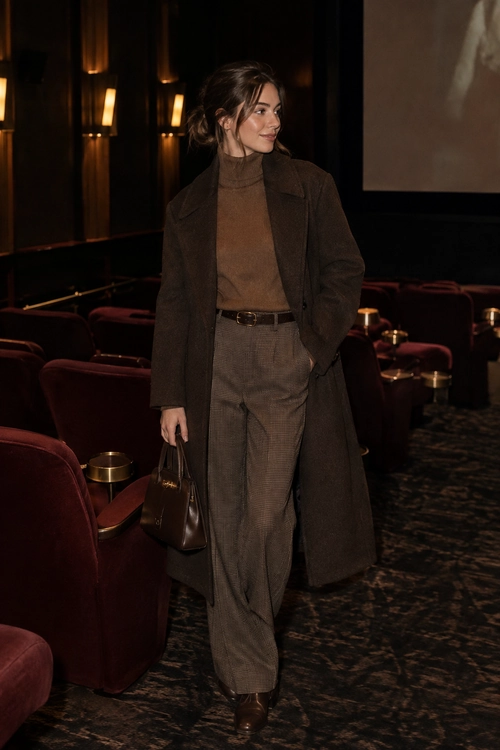 A woman wears a long brown coat over a taupe turtleneck, checked trousers, brown ankle boots, and carries a brown leather handbag in a cinema.
