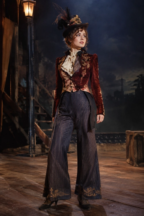 A woman wears a burgundy velvet embroidered jacket, ruffled cream blouse, pinstriped wide-leg trousers, black boots, and a feathered top hat.